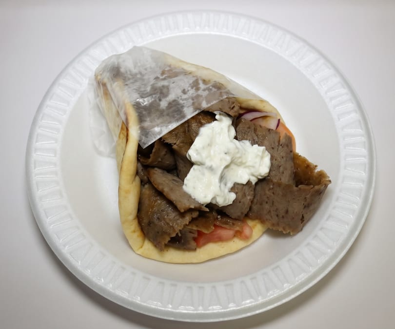 A gyro with meat, tomatoes, and tzatziki sauce on a white styrofoam plate.