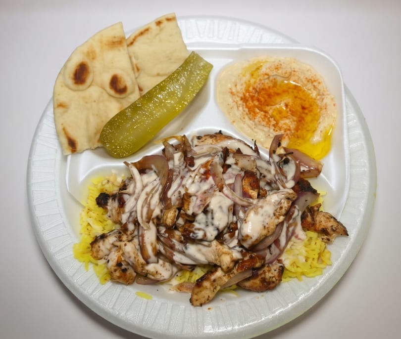 Chicken shawarma plate with rice, hummus, pita bread, and a pickle spear on a white plate.