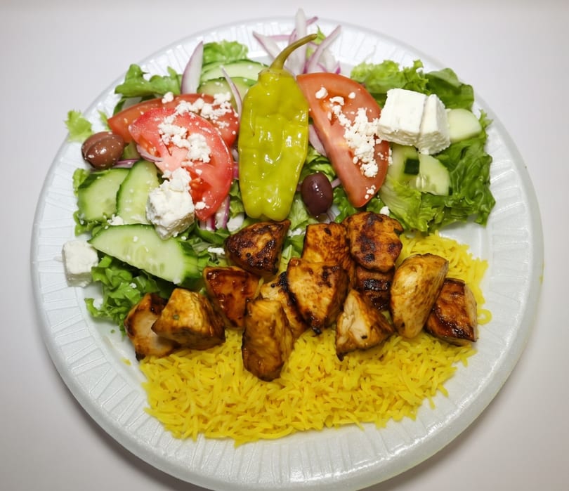 Chicken tikka with yellow rice, Greek salad, and a pepperoncini on a white plate.